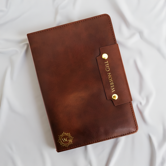The Webson Gill Leather Writing Notebook features a brown leather cover with gold emblem and branding, ideal for fountain pen enthusiasts, shown resting on white fabric.
