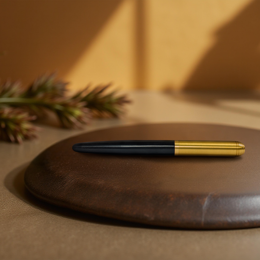 The Webson Gill 'Captain Edition' Premium Fountain Pen in black wood with a brass cap rests on a round brown leather pad, set against blurred greenery in the background.