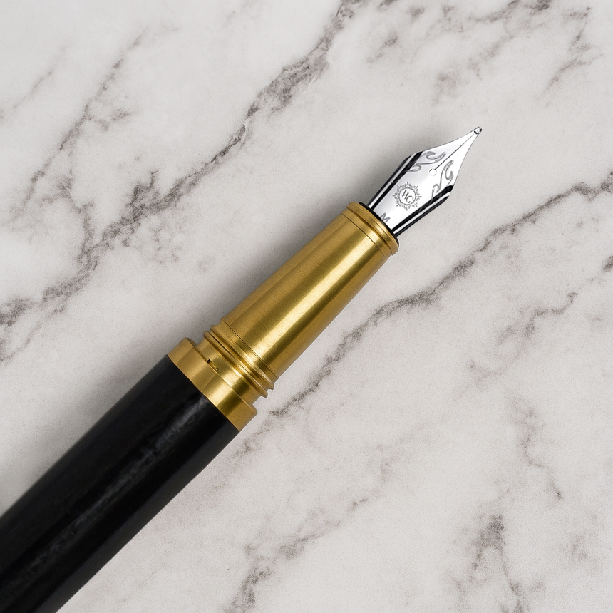 The Webson Gill 'Captain Edition' Premium Fountain Pen - Black Wood, featuring a gold and silver nib and a brass cap, rests elegantly on a white marble surface.