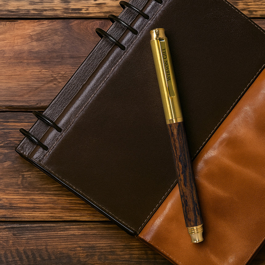 The Webson Gill 'Prestige Edition' Premium Fountain Pen with an oak wood barrel rests on a brown leather notebook atop a wooden table.