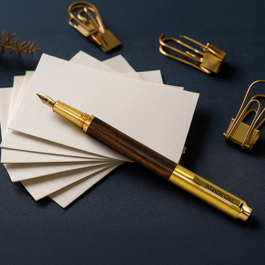 The Webson Gill 'Prestige Edition' Premium Fountain Pen with an oak wood barrel rests on cream cards, accented by gold paper clips on a dark surface.
