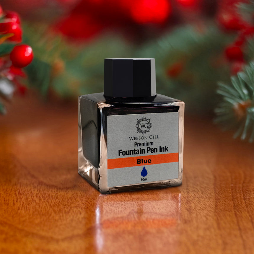 A bottle of Webson Gill's 'Ink in a Bottle' Fountain Pen Refill in blue sits on a wooden surface, complemented by festive red berries in the background.