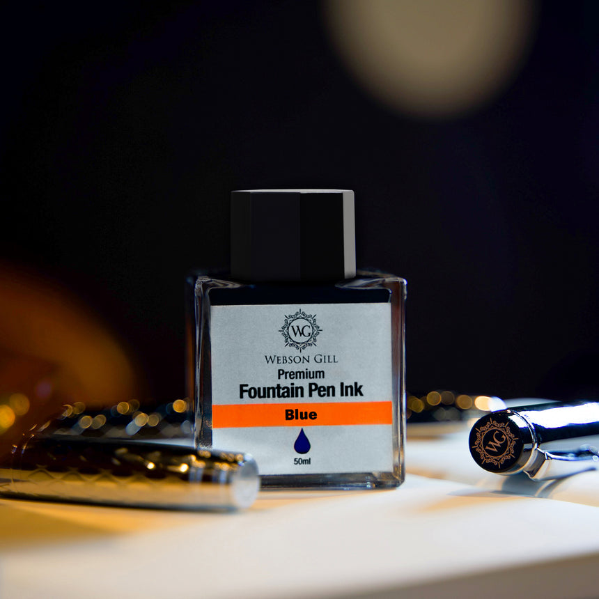 A bottle of Webson Gill 'Ink in a Bottle' Fountain Pen Refill (blue ink, fits all fountain pens) sits with two silver pens on a dark surface.
