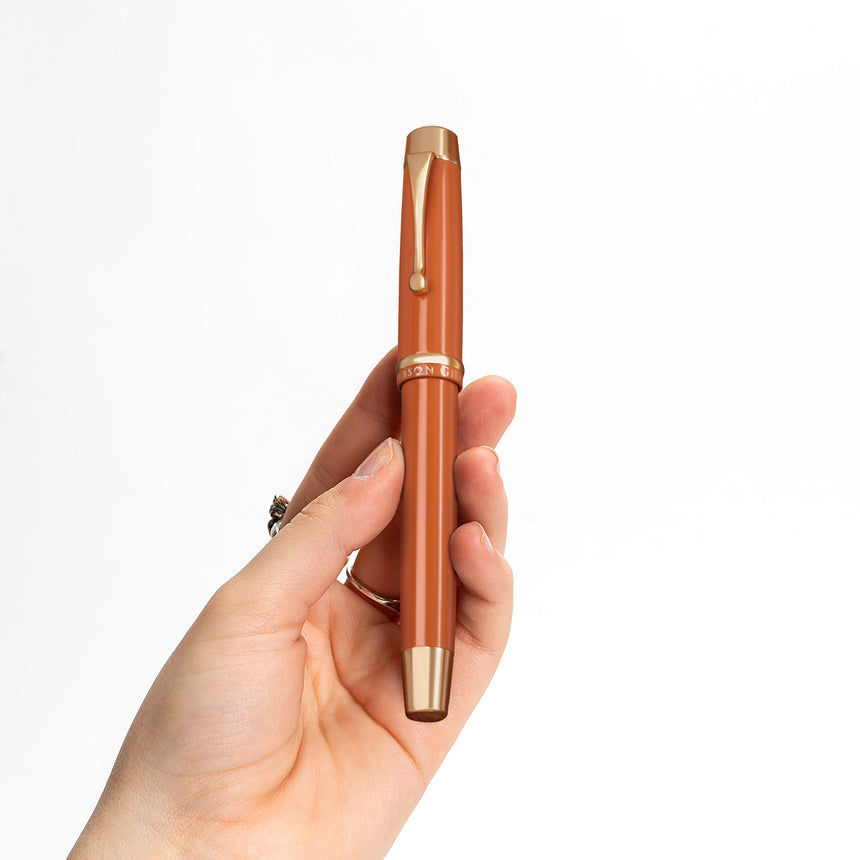 A hand holds The Muse Fountain Pen by Webson Gill, featuring an orange body and a gold-plated nib, against a white background.