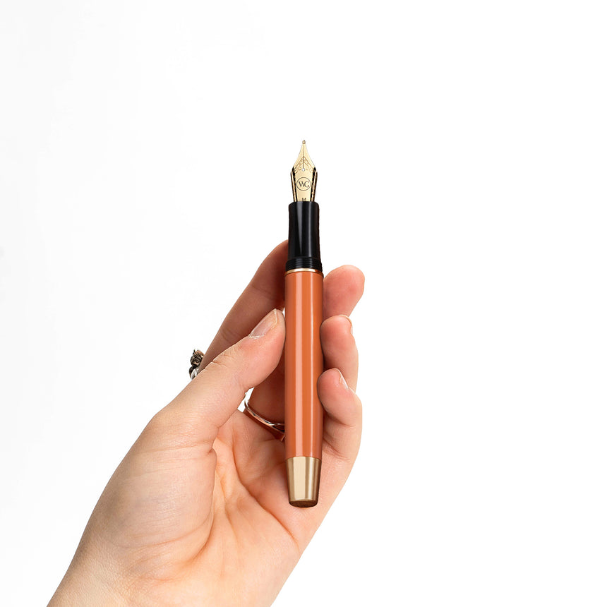 A hand holding The Muse Fountain Pen by Webson Gill, featuring an orange body, gold-plated nib, and ergonomic design, set against a plain white background.