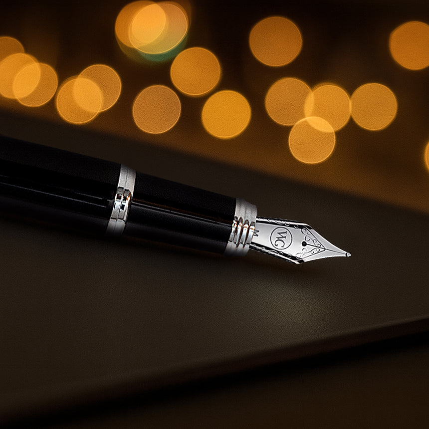 The Webson Gill 'The Mogul' Premium Fountain Pen, featuring a stainless steel nib, rests on a surface with blurred golden lights glowing in the background.