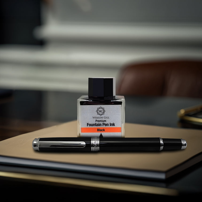 A Webson Gill 'The Mogul' Premium Fountain Pen with a stainless steel nib and black ink bottle rest elegantly on a closed notebook.