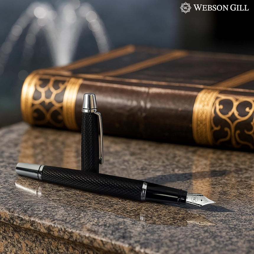 A 'Show Up' Everyday Fountain Pen by WG, featuring a black body and stainless steel medium nib, rests on a stone surface next to an ornate book, with the WG logo visible in the corner.