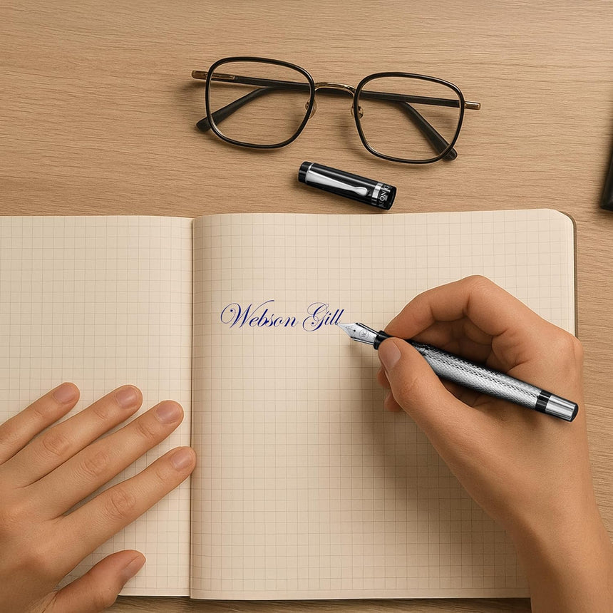Someone writes "Webson Gill" in a grid notebook using WG’s ‘The Raider’ Everyday Fountain Pen, which has a black and silver design and a stainless steel medium nib; glasses lie nearby on the desk.
