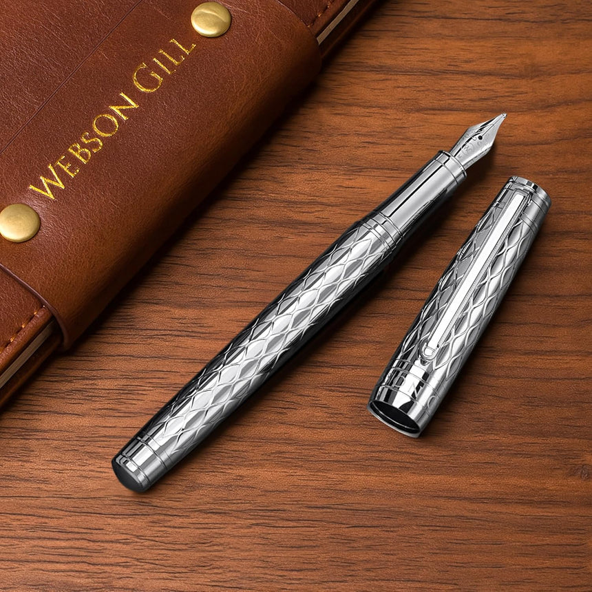 The WG ‘The Mirror’ Everyday Fountain Pen, with a silver metal finish, patterned cap and barrel, and stainless steel nib, rests on a wooden desk next to a brown leather case.