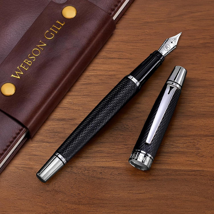 The WG 'Show Up' Everyday Fountain Pen in black and silver with a stainless steel medium nib rests uncapped next to a brown leather notebook on a wooden desk.