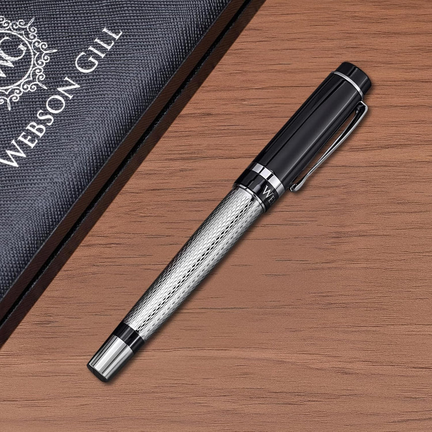 A black and silver ‘The Raider’ Everyday Fountain Pen by WG, featuring a stainless steel medium nib, rests on a wooden surface beside a dark textured WG box.