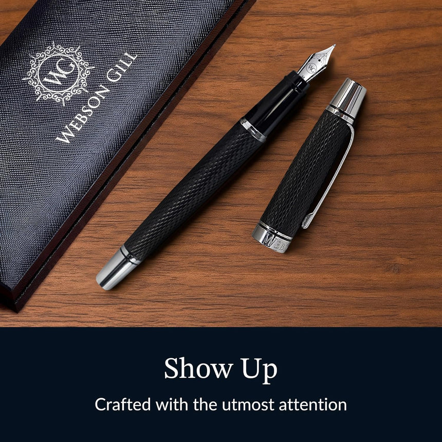 A black WG 'Show Up' Everyday Fountain Pen with a diamond guilloché pattern, cap off, rests beside its gift box on a wooden surface. Text: "Show Up.