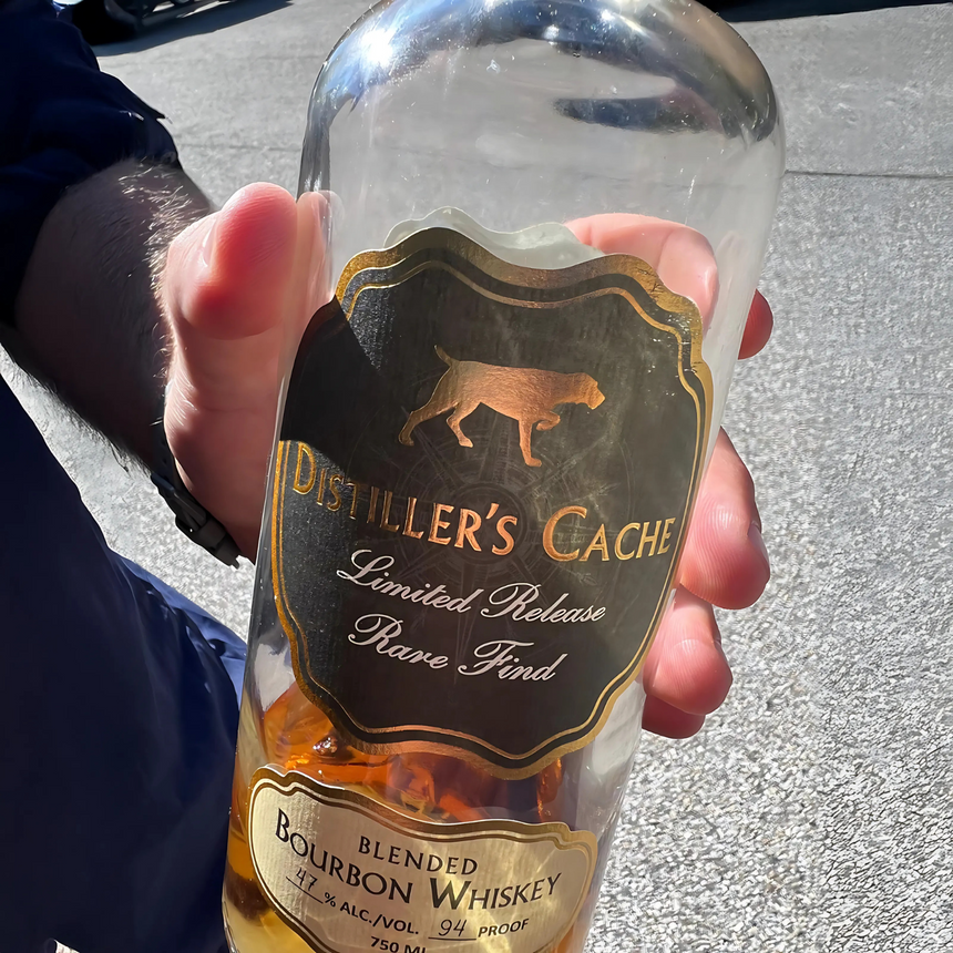 A hand holds a bottle of Distiller’s Cache Blended Bourbon Whiskey outdoors in sunlight, next to a Webson Gill 'Bourbon Barrel' Premium Fountain Pen crafted from reclaimed oak wood.