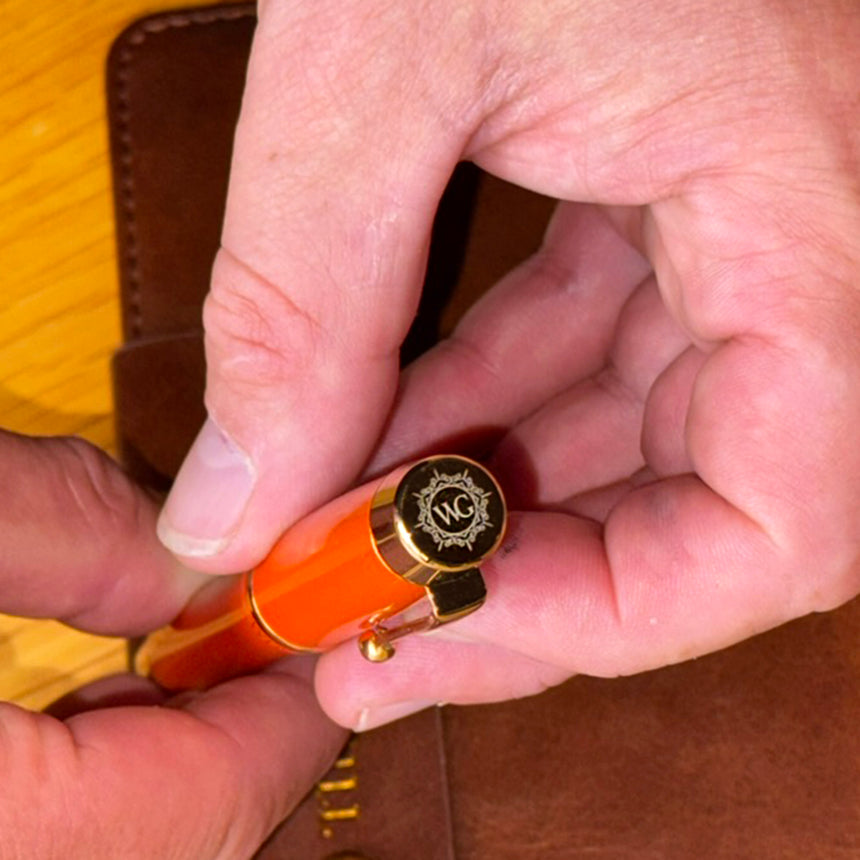 A hand holds The Muse Fountain Pen by Webson Gill, featuring an orange body, gold "WC" emblem, and ergonomic design near a brown leather case on a wooden surface.