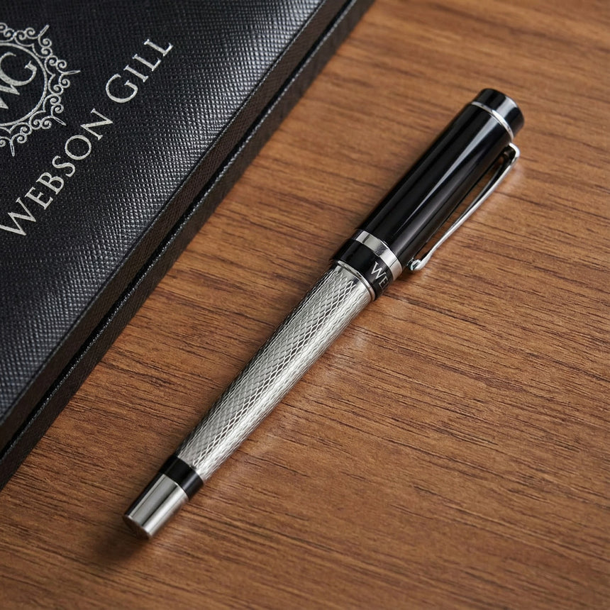 The WG ‘The Raider’ Everyday Fountain Pen, featuring a black and silver design with a stainless steel medium nib, rests on a wooden surface beside a book.