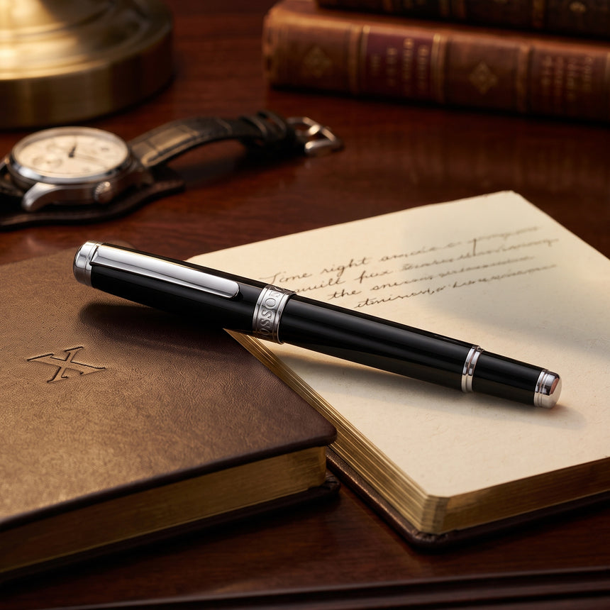A sleek black 'The Mogul' Premium Ballpoint Pen by Webson Gill with a smooth ink flow rests on an open notebook, accompanied by a closed book, watch, and lamp in the background.