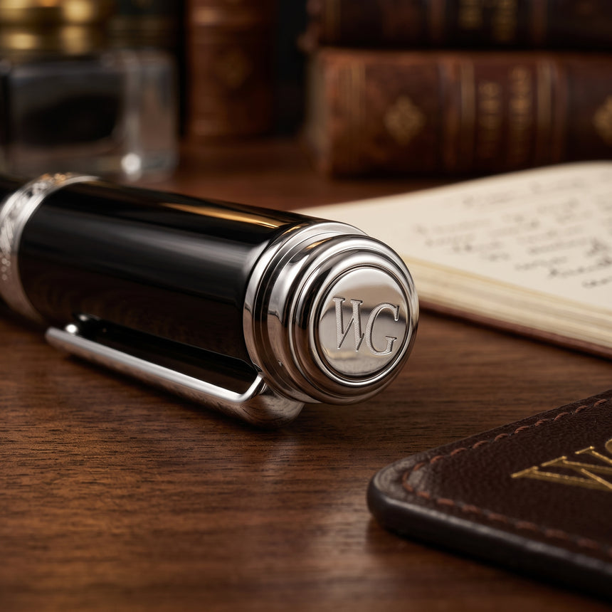 A black and silver 'The Mogul' Premium Ballpoint Pen by Webson Gill, engraved with WG and renowned for its smooth ink flow, rests on a wooden desk beside a notebook and books.