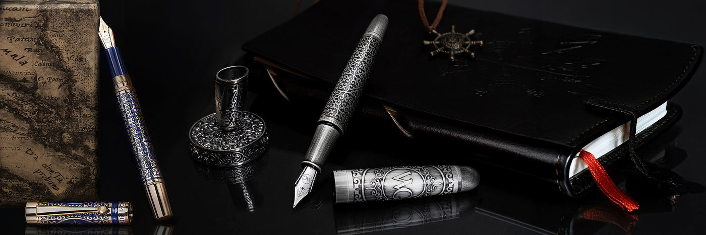 Two ornate fountain pens and a black leather journal with a red ribbon on a reflective black surface.