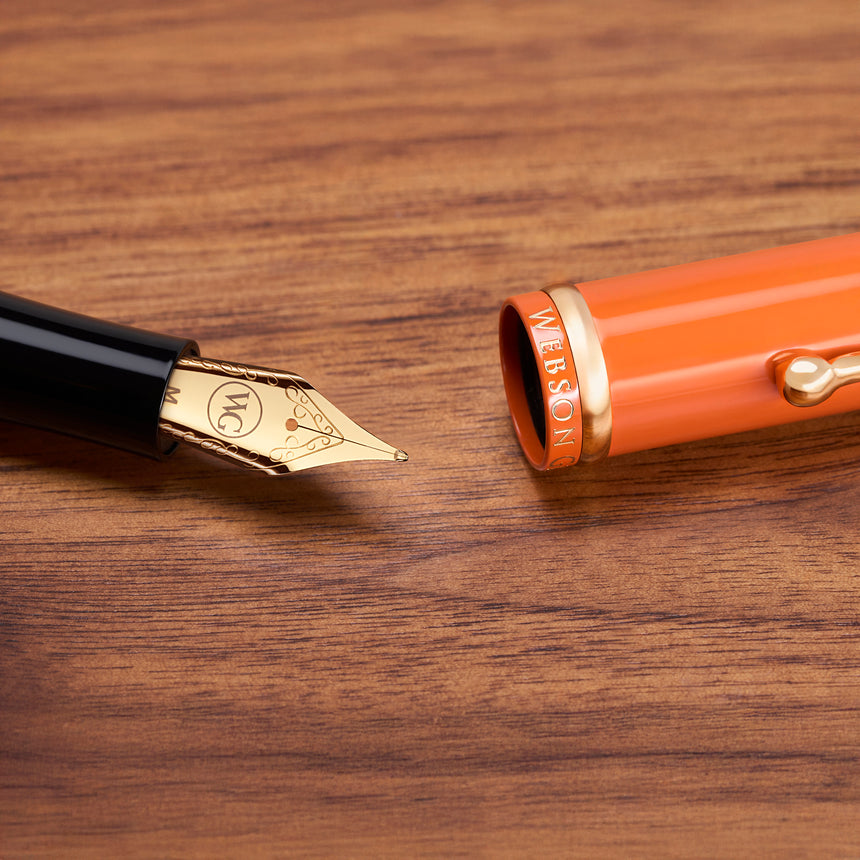 The Muse Fountain Pen by Webson Gill features a gold-plated nib resting beside its orange cap on a wooden surface, highlighting its elegant and ergonomic design.