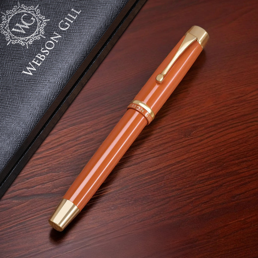 A gold and orange Muse Fountain Pen by Webson Gill with an ergonomic design rests on a wooden surface beside its black case.