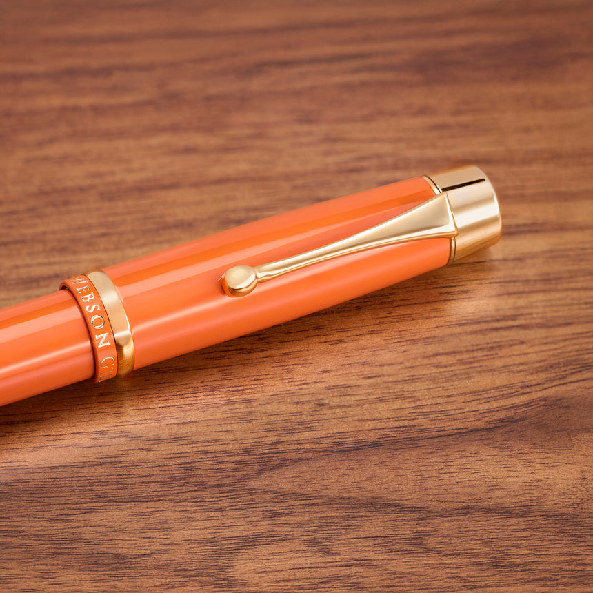 The Muse Fountain Pen by Webson Gill, featuring an orange body, gold trim, and a gold-plated nib, rests on a wooden surface and highlights its ergonomic design.