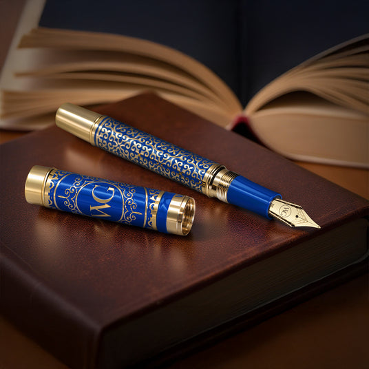 The Regal Fountain Pen by Webson Gill, featuring elegant blue and gold brass filigree, rests on a closed brown book with an open book behind—a stunning piece for any fountain pen collector.