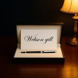 A Webson Gill card and The Mogul Fountain Pen, known for its comfortable grip, are displayed in a gift box on a wooden table beside a lit lamp.