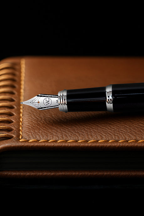 A black fountain pen rests on a brown leather-bound notebook with detailed stitching.