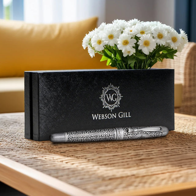 The Webson Gill Admiral Fountain Pen, featuring an antique silver finish, rests in a black branded box on a table, highlighting its elegant design and craftsmanship with white flowers in the background.