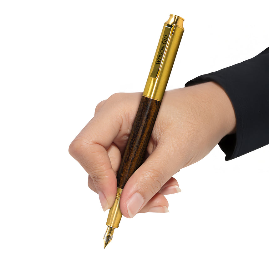 A hand holds the Webson Gill Prestige Edition Fountain Pen, featuring a gold and wood finish with an iridium nib, ready to write against a white background.