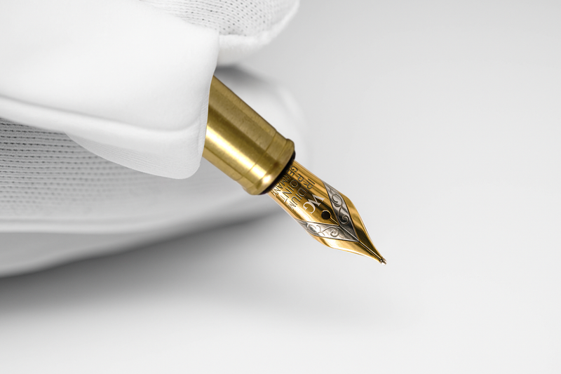 A gloved hand holds a gold fountain pen with an ornate engraved nib against a white background.