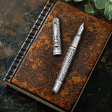 The Admiral Fountain Pen by Webson Gill, featuring an antique silver finish, rests atop a vintage brown leather-bound notebook, highlighting refined craftsmanship.