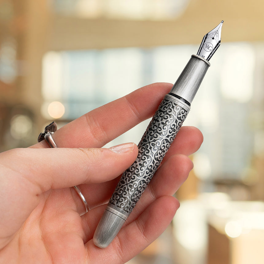 A hand holds the Webson Gill Admiral Fountain Pen, a luxurious writing instrument with an ornate engraved silver barrel, in a softly blurred indoor setting.
