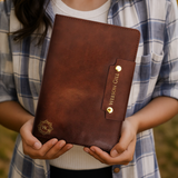 A person in a plaid shirt holds a brown Leather Writing Notebook by Webson Gill with gold accents.