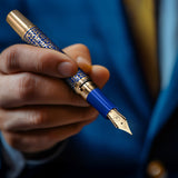A hand holds The Regal Fountain Pen by Webson Gill, featuring elegant blue and gold brass filigree—an ideal choice for fountain pen collectors—set against a blurred blue suit background.