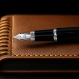 The Mogul Fountain Pen by Webson Gill, featuring a glossy black resin finish, rests atop a brown leather notebook with decorative stitching.