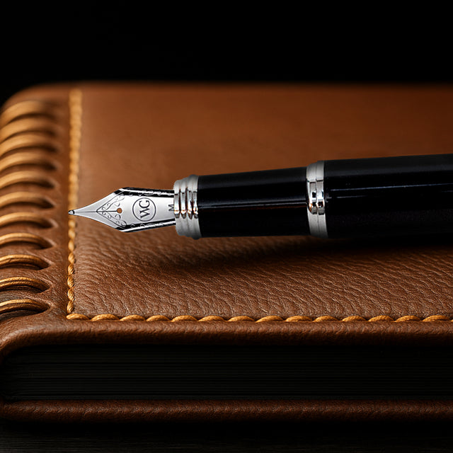 The Mogul Fountain Pen by Webson Gill, featuring a glossy black resin finish, rests atop a brown leather notebook with decorative stitching.
