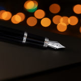 The Mogul Fountain Pen by Webson Gill, featuring a stainless steel nib and black finish, rests on a dark surface with blurred orange lights glowing in the background.
