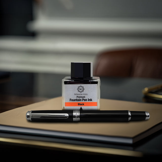 The Mogul Fountain Pen by Webson Gill, featuring a comfortable grip and stainless steel nib, rests with its black ink bottle on a closed notebook atop the desk.