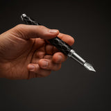 A hand holds WG’s ‘The Weaver’ Everyday Fountain Pen, featuring a black and silver lattice design and a stainless steel medium nib, poised against a dark background.