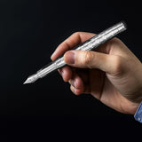 A hand holds WG’s ‘The Mirror’ Everyday Fountain Pen—a silver, patterned metal pen with a stainless steel nib—against a dark background.
