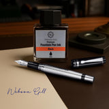 ‘The Raider’ Everyday Fountain Pen by WG, featuring a stainless steel medium nib, rests next to a black WG ink bottle on a desk with paper and the pen cap.