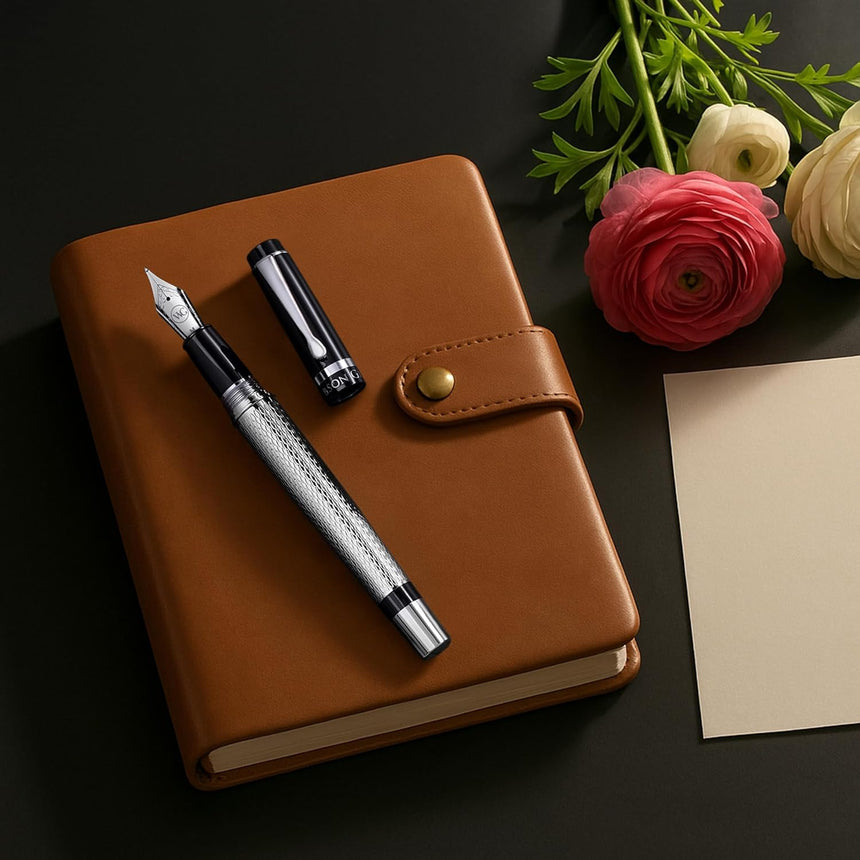 A brown leather notebook with WG’s ‘The Raider’ Everyday Fountain Pen, featuring a black and silver body and stainless steel medium nib, is placed beside flowers and blank paper on a dark surface.