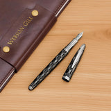 The WG ‘The Weaver’ Everyday Fountain Pen in black and silver lattice, with a matching cap beside a brown WEBSON GILL leather folder, features a stainless steel medium nib for smooth writing.