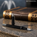 A 'Show Up' Everyday Fountain Pen by WG, featuring a black body and stainless steel medium nib, rests on a stone surface next to an ornate book, with the WG logo visible in the corner.