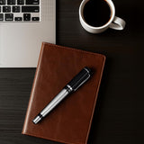 The WG ‘The Raider’ Everyday Fountain Pen, featuring a black and silver finish and a stainless steel medium nib, rests on a brown notebook beside a laptop and a cup of black coffee on a dark desk.