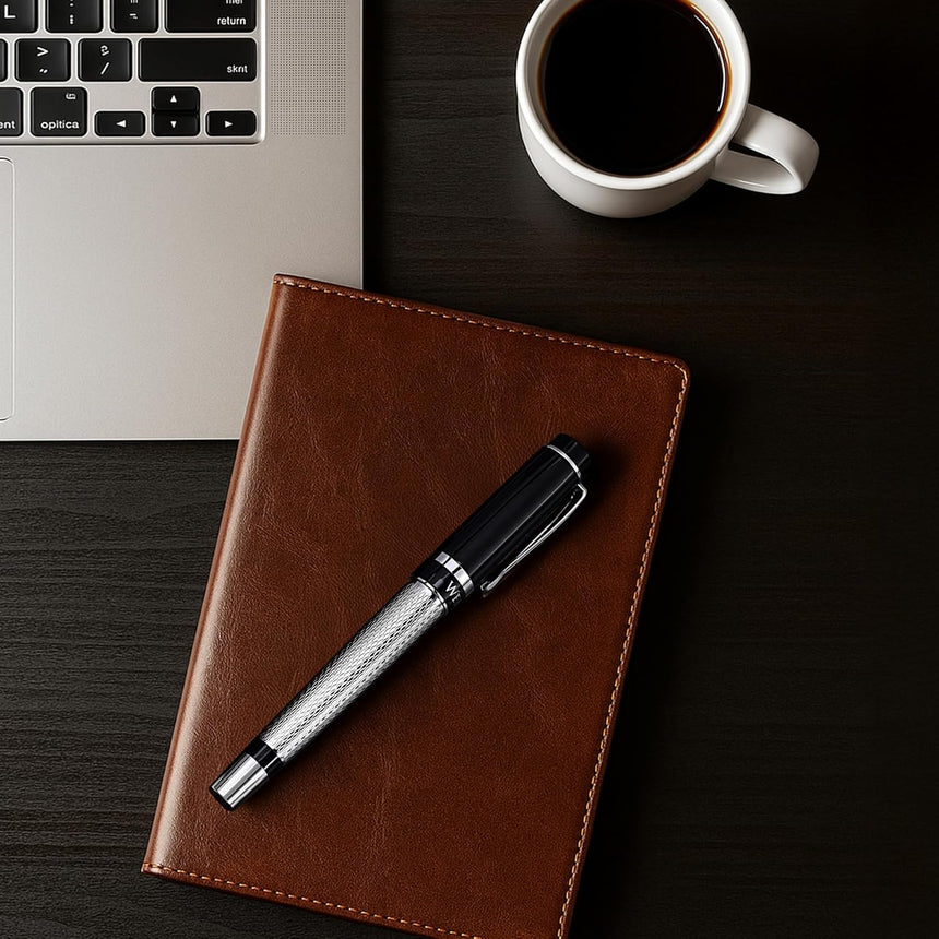 The WG ‘The Raider’ Everyday Fountain Pen, featuring a black and silver finish and a stainless steel medium nib, rests on a brown notebook beside a laptop and a cup of black coffee on a dark desk.