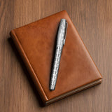 The WG ‘The Mirror’ Everyday Fountain Pen with a stainless steel nib rests on a closed brown leather notebook on a wooden surface.