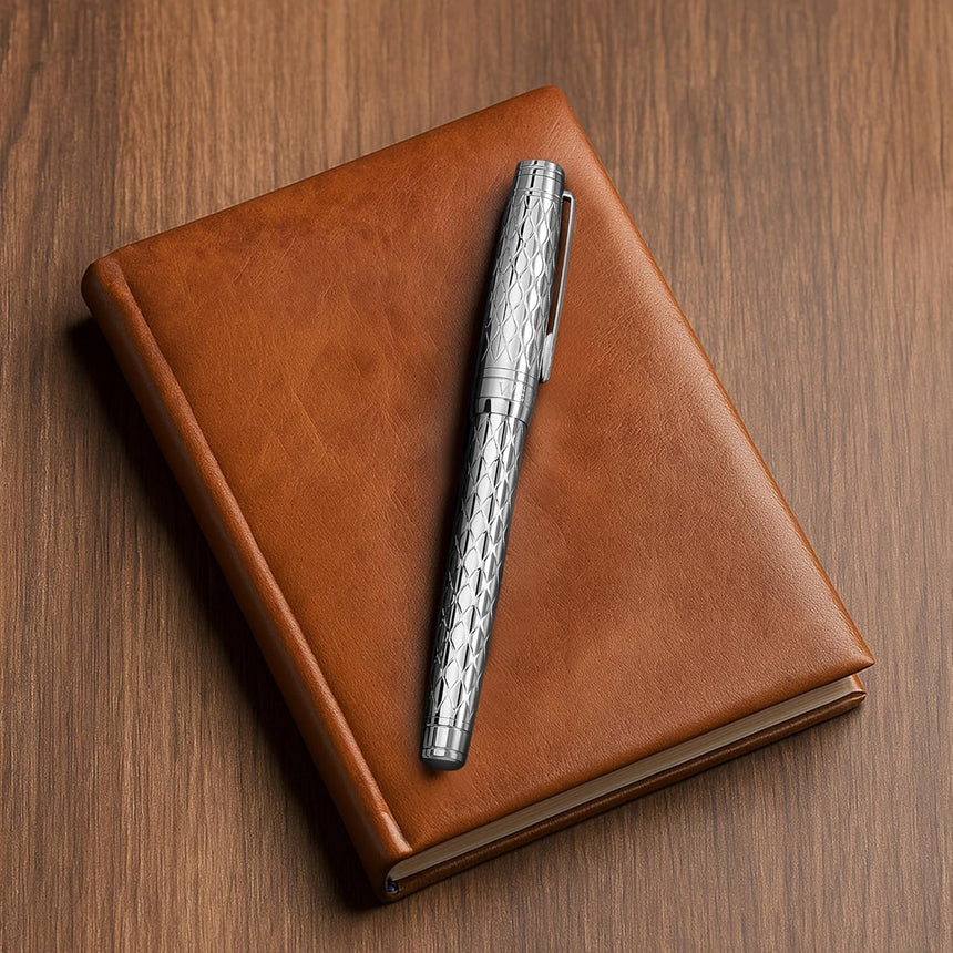 The WG ‘The Mirror’ Everyday Fountain Pen with a stainless steel nib rests on a closed brown leather notebook on a wooden surface.